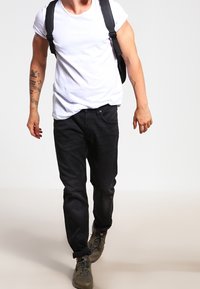 Vit t-shirt med kort ärm, svarta jeans, grå sneakers med texturerade sulor och en svart ryggsäck med justerbara remmar.