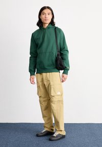 Grüner Kapuzenpullover, beige Cargo-Hosen mit Taschen, schwarze Umhängetasche und dunkle Schuhe. Einfaches Design, lässige Passform, matte Textur.