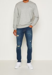 Šedá mikina s žebrovanými manžetami a lemem, v kombinaci s tmavomodrými roztrhanými skinny džíny a bílými teniskami. Ležérní outfit s minimálním logem.