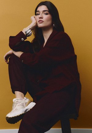 Mujer con cabello largo y oscuro posando sentada, vestida con un conjunto burdeos oscuro, zapatillas beige y una pulsera ancha plateada, sobre un fondo amarillo mostaza.