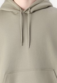 Olive-green hoodie made of soft fabric, featuring a front pocket, adjustable drawstring with silver tips, and ribbed hemline.