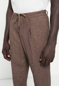 Brown houndstooth trousers made of textured fabric, featuring a drawstring waist, pleats, and side pockets.