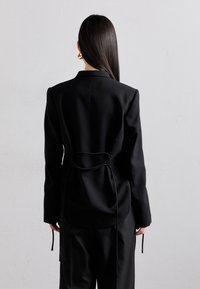 Schwarze Blazer mit strukturiertem Design, langen Ärmeln und einem Bindeband im Rücken. Der Stoff wirkt glatt und maßgeschneidert.