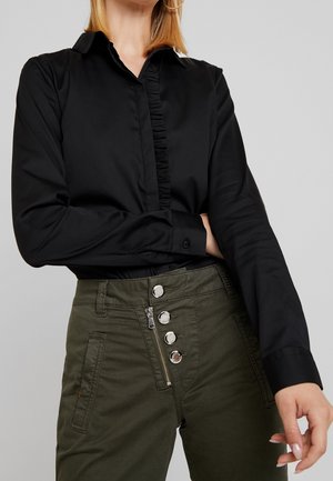 Maglietta nera a maniche lunghe con un davanti arricciato, abbinata a pantaloni verde oliva a vita alta con più bottoni argentati e chiusura con zip.
