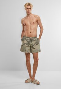 Pantaloni da bagno corti di un leggero beige con un sottile motivo mimetico. Vita elasticizzata con tasche laterali, presentati con sandali minimalisti.