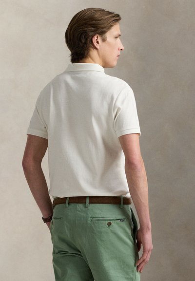 Mann i hvit kortermet poloshirt og grønne bukser med brunt belte, stående sideveis mot en ensfarget beige bakgrunn.