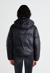 Schwarze Steppjacke mit Kapuze, die gepolsterte Abschnitte und eine glänzende, glatte Oberfläche aufweist. Darüber werden dunkelglänzende Leggings getragen.