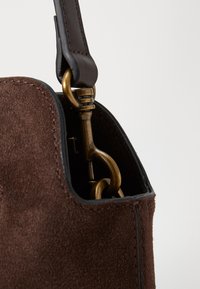 Braune Wildledertasche mit einem Ledertragegurt, ausgestattet mit einem Messingverschluss und schwarzen Kanten. Weiche Textur mit sichtbaren Steppdetails.