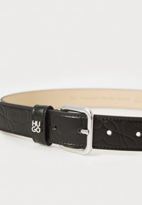 Ceinture en cuir noir avec un motif embossé, boucle en métal argenté et accent de marque. Dispose de plusieurs trous pour un ajustement de taille.