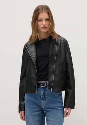 Junge Frau trägt eine schwarze Lederjacke über einem schwarzen Shirt, kombiniert mit blauen Jeans und einem schwarzen Gürtel vor einem schlichten Hintergrund.