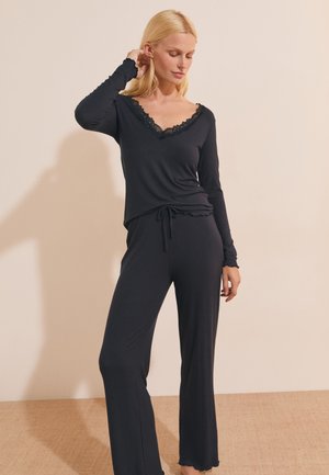 Vrouw met blond haar die een zwarte top met lange mouwen en kanten halslijn draagt, en bijpassende zwarte wijde broek, staand tegen een beige muur.