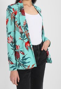Marynarka w kwiaty w turkusowym kolorze, zdobiona czerwonymi i różowymi kwiatami, z dwiema przednimi kieszeniami i luźnym krojem, zestawiona z białą bluzką i czarnymi dżinsami.