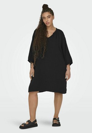 Schwarzes, knielanges Kleid mit kurzen, lockeren Ärmeln und einem V-Ausschnitt. Gewebte Struktur, akzentuiert durch eine fließende Silhouette. Schwarze Sandalen runden den Look ab.