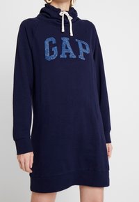 Granatowa sukienka bluza wykonana z miękkiego materiału, z wysokim kołnierzem i białym sznurkiem. Na froncie widoczny napis "GAP" w niebieskim brokatowym kolorze.