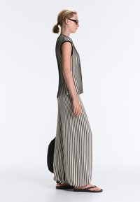 Femme avec des lunettes de soleil porte un haut sans manches rayé vertical noir et blanc et un pantalon large assorti, portant un sac noir et des sandales.