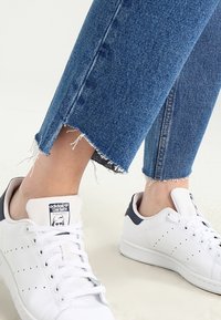 Vita lädersneakers Adidas Stan Smith med marinblå detaljer, kombinerade med blå jeans med fransiga kanter.