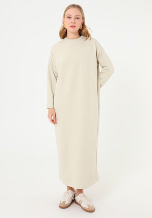 Kvinde med skulderlangt blondt hår iført beige midi-kjole med lange ærmer og hvide sneakers, stående mod hvid baggrund.