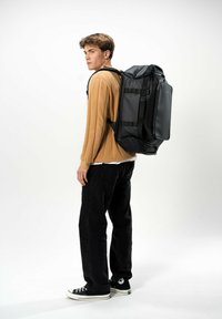 Grande zaino nero con spallacci imbottiti, design elegante, molteplici scomparti e manici laterali, indossato da una persona con un maglione beige e pantaloni neri.