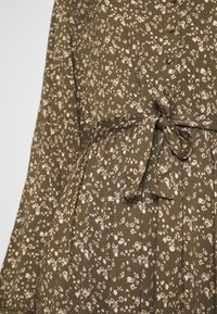 Abito a fiori in verde oliva con fiori crema. Presenta una vita con lacci, pieghe e dettagli con bottoni lungo la parte anteriore. Tessuto dalla consistenza morbida.