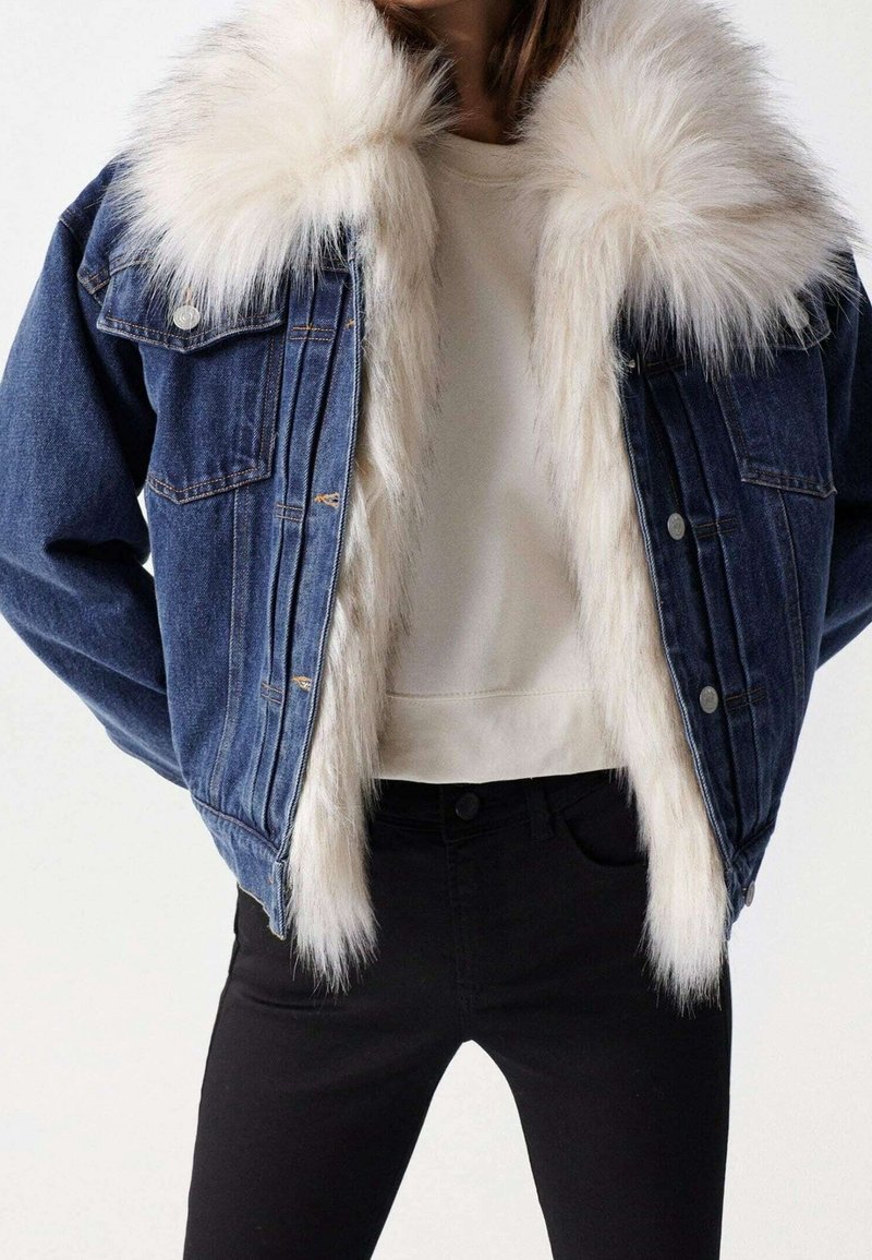 Jeansjacka med stor vit syntetisk pälskrage och innerfoder, som bärs över en ljusbeige topp och svarta byxor.