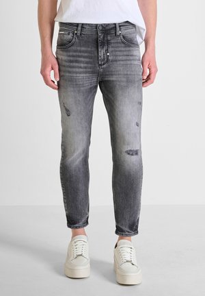 Jeans skinny gris desgastados con detalles de decoloración y parches; cuenta con cinco bolsillos y cierre de botón. Llevados con zapatillas blancas.