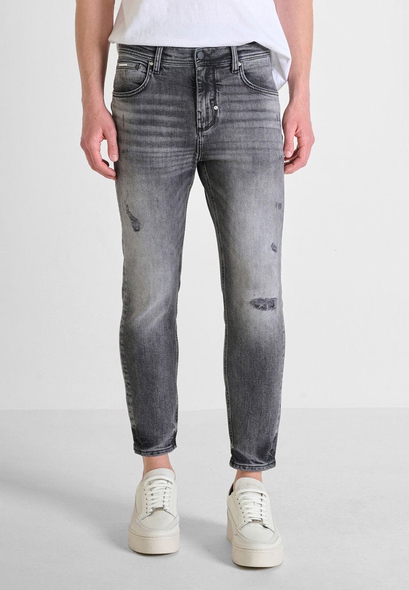 Jeans skinny gris desgastados con detalles de decoloración y parches; cuenta con cinco bolsillos y cierre de botón. Llevados con zapatillas blancas.