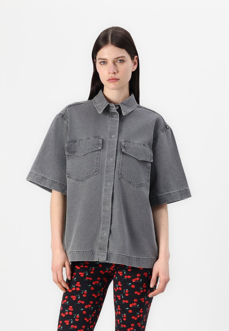 Kortermet, grå denimskjorte med krave og to brystlommer. Brukt over mønstrede svarte leggings med kirsebærdesign.