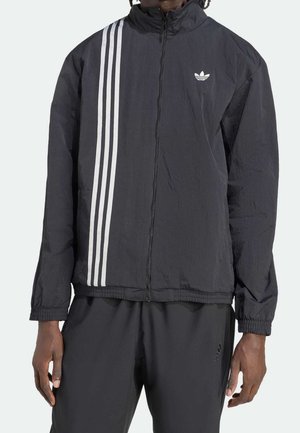 Veste zippée noire Adidas avec trois bandes blanches sur le côté gauche et logo Adidas sur la poitrine, portée avec un pantalon Adidas noir.