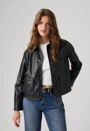 Femme portant une veste en cuir noire sur un crop top blanc et un jean bleu avec une ceinture noire, debout devant un fond clair uni.
