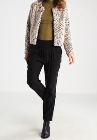 Faux-Fur-Jacke in Creme mit schwarzen Leopardenflecken, kombiniert mit einem grünen Oberteil und schwarzen plissierten Hosen. Abgerundet mit schwarzen Knöchelstiefeln.