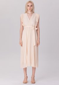 Robe plissée beige clair avec des manches volantes, un col en V et une ceinture nouée à la taille, présentant un design fluide et une texture douce.