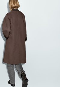 Cappotto lungo marrone con collo, caratterizzato da maniche larghe e una linea dritta. Abbinato a jeans grigi e stivali neri.