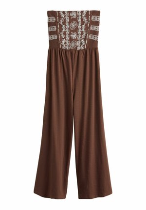 Junge Frau mit lockerem dunklem Haar, die ein trägerloses, gemustertes braunes Kleid trägt, steht nahe am Wasser unter klarem Himmel mit Sonnenlicht.