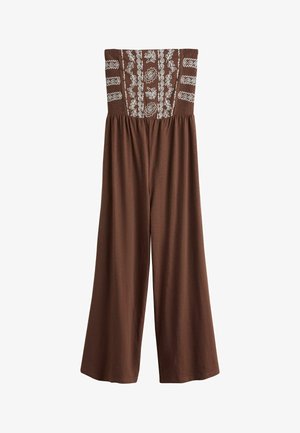 Jeune femme aux cheveux foncés détachés, portant une robe marron à motifs sans bretelles, debout près de l'eau sous un ciel dégagé avec la lumière du soleil.