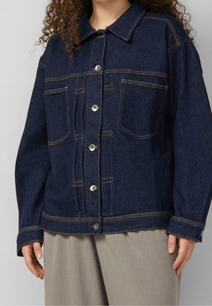 Personne portant une veste en jean bleu foncé avec des boutons à l'avant et deux poches poitrine, associée à un pantalon beige clair à jambes larges.