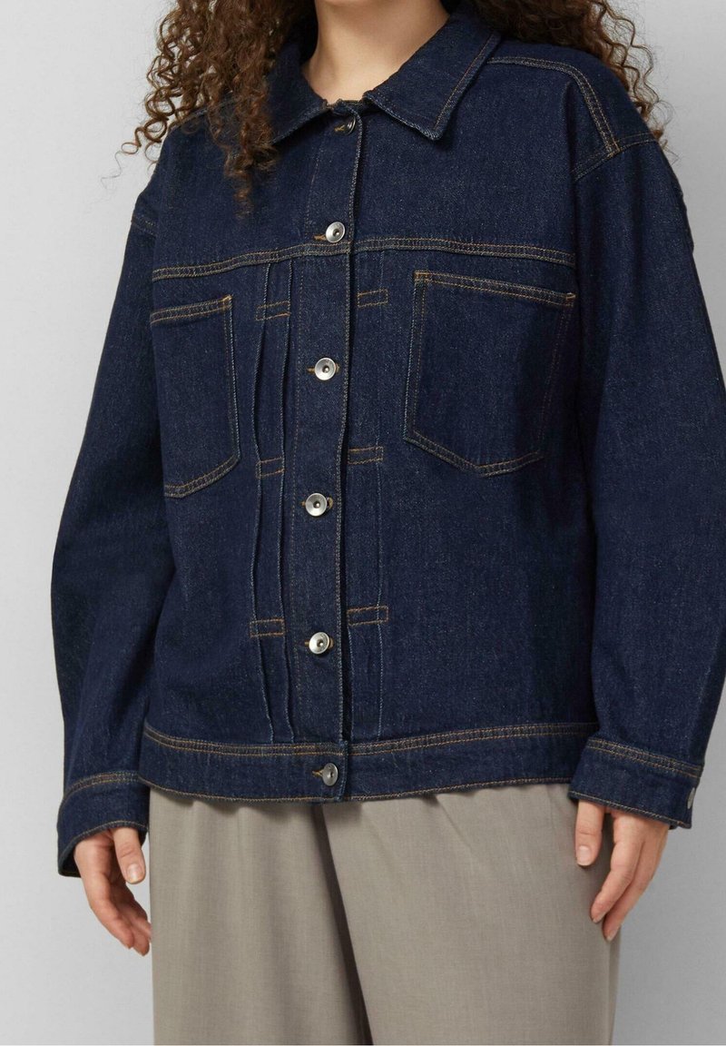 Personne portant une veste en jean bleu foncé avec des boutons à l'avant et deux poches poitrine, associée à un pantalon beige clair à jambes larges.