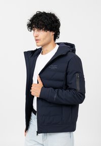 Marineblaue Steppjacke mit Kapuze, Reißverschlussfront und Seitentaschen. Mit einem Logo auf der Brust und einer Reißverschlusstasche am Arm. Glatte Textur.