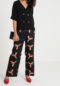 Černá dvouřadá halenka s vyhrnutými rukávy, v kombinaci se širokými kalhotami s oranžovo-modrými vzory ptáků, doplněná červenými botami a psaníčkem.