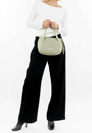 Vrouw in een wit top met lange mouwen, zwarte wijde broekspijpen, zwarte laarzen met hak, en ze houdt een kleine lichtgroene geperforeerde handtas met sleutelhanger vast.