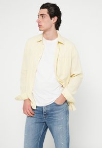 Ανοιχτό κίτρινο, ριγέ πουκάμισο με κουμπιά από ύφασμα, φορεμένο πάνω από λευκό Τ-shirt, συνδυασμένο με φθαρμένο μπλε τζιν. Απλή λεπτομέρεια τσέπης.