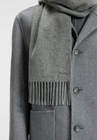 Sciarpa di lana grigia con dettagli frangiati, appoggiata su un cappotto grigio abbinato con bottoni arrotondati e una tasca frontale.