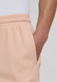 Licht perzikkleurige, gestructureerde shorts met elastische tailleband en zijzakken, gedeeltelijk zichtbaar met een hand in een zak. Het materiaal lijkt zacht en ademend.