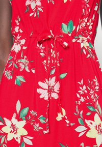 Zblízka červené květinové šaty s bílými, žlutými a zelenými květinovými vzory a uvázaným látkovým páskem v pase.