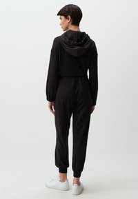 Zwarte jumpsuit met capuchon, lange mouwen, elastische tailleband en manchetten aan de enkels, gecombineerd met witte sneakers. Gladde textuur en een relaxte pasvorm.
