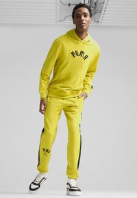 Gul huvtröja och matchande joggers med svarta accenter, med en framficka och logotyp. Sneakers har vit och svart detaljering.