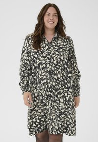 Vrouw met lang bruin haar in een zwart-wit gestreepte blousejurk met lange mouwen, die glimlacht en tegen een effen achtergrond staat.