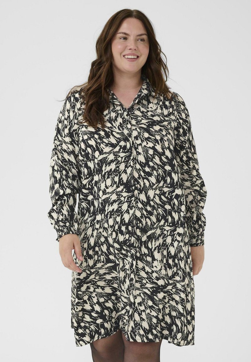 Vrouw met lang bruin haar in een zwart-wit gestreepte blousejurk met lange mouwen, die glimlacht en tegen een effen achtergrond staat.