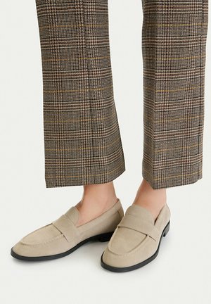 Beige Wildleder-Loafers mit schwarzen Sohlen, getragen mit braunen karierten, gekürzten Hosen barfuß vor einem neutralen Hintergrund.