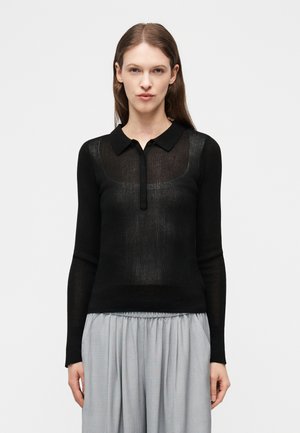 Γυναίκα με μακριά καστανά μαλλιά που φοράει μαύρο διάφανο μπλουζάκι μακρυμάνικο με γιακά και γκρι πλισέ φούστα, στέκεται μπροστά σε απλό φόντο.