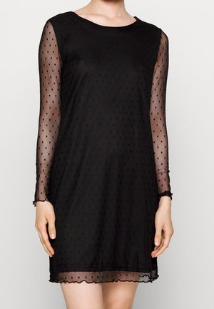 Vrouw draagt een zwarte knie-lange jurk met doorschijnende lange mouwen en kleine stippenpatronen, staand tegen een effen achtergrond.